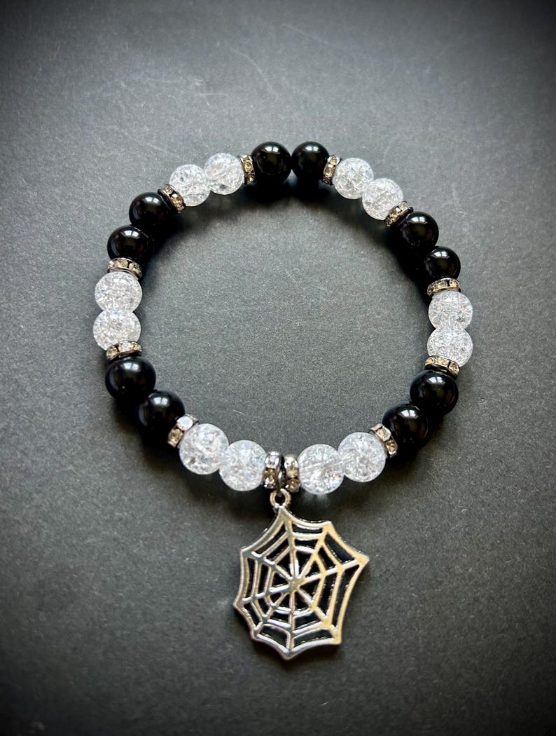 May include: A black and white beaded bracelet with a silver spiderweb charm. The bracelet features alternating black and clear, crackled beads, with small silver accents. The spiderweb charm hangs from the center of the bracelet.