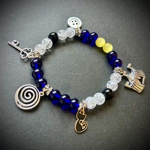 May include: A beaded bracelet with blue, clear, black, and yellow beads. The bracelet features silver charms including a key, a spiral, a cat, a button, and a crown. The bracelet is on a dark background.