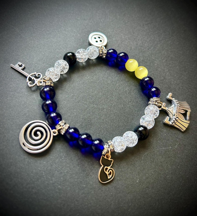 May include: A beaded bracelet with blue, clear, black, and yellow beads. The bracelet features silver charms including a key, a spiral, a cat, a button, and a crown. The bracelet is on a dark background.