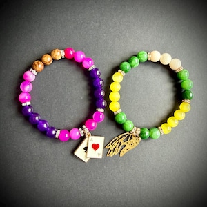 May include: Two beaded stretch bracelets. The left bracelet features pink, purple, and brown beads with a gold-tone charm of two playing cards, one showing an ace of hearts. The right bracelet features green, yellow, and cream beads with a gold-tone hand charm.