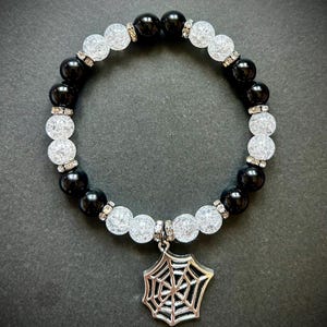 May include: A beaded bracelet featuring alternating black and white, textured beads. The bracelet has a silver spiderweb charm. The beads are accented with small, silver-colored spacers. The bracelet is displayed on a dark surface.