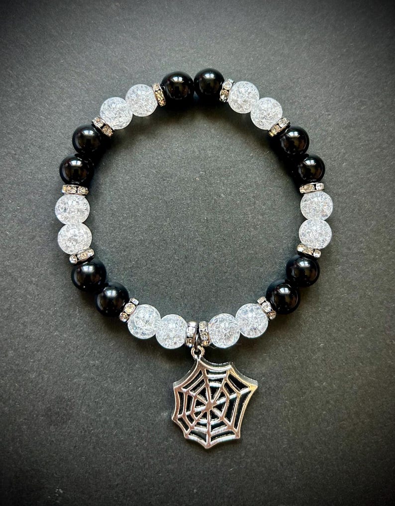 May include: A beaded bracelet featuring alternating black and white, textured beads. The bracelet has a silver spiderweb charm. The beads are accented with small, silver-colored spacers. The bracelet is displayed on a dark surface.