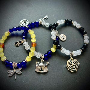 May include: Three beaded bracelets with charms. One bracelet has blue and yellow beads with a dragonfly charm. Another has blue, yellow, and clear beads with a circus tent charm. The third has black and clear beads with a spiderweb charm.