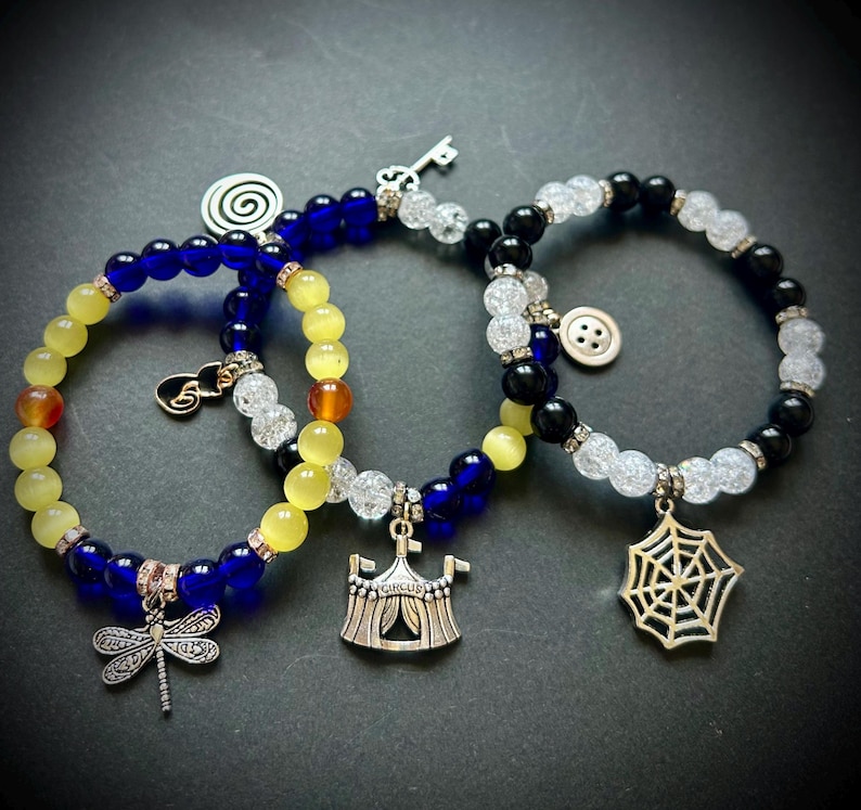 May include: Three beaded bracelets with charms. One bracelet has blue and yellow beads with a dragonfly charm. Another has blue, yellow, and clear beads with a circus tent charm. The third has black and clear beads with a spiderweb charm.
