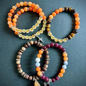 May include: A selection of beaded bracelets in shades of orange, yellow, brown, and purple. Some bracelets have decorative charms and sparkling details. The bracelets are displayed on a dark background.