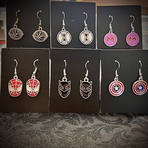 Könnte beinhalten: Eine Sammlung von silberfarbenen Ohrhängern mit verschiedenen Superhelden-Designs. Die Ohrringe zeigen Bilder eines Schildes, einer Spider-Man-Maske, eines schwarzen Panthers und anderer Comic-Symbole. Jedes Paar wird auf einer schwarzen Karte präsentiert.
