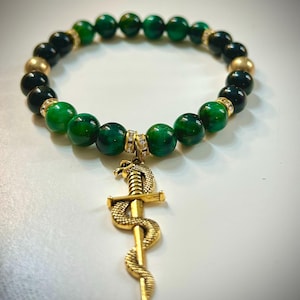 Könnte beinhalten: Ein Armband mit grünen und schwarzen Perlen, unterbrochen von goldfarbenen Perlen. Ein goldfarbener Anhänger mit einer Schlange, die sich um ein Schwert windet, hängt am Armband. Modeschmuck.