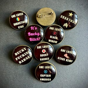 Peut inclure: Ensemble de badges ronds en émail brun foncé avec des textes blancs et roses. Les badges affichent des phrases comme "She Loves Bucky Barnes" et "It's Bucky, Bitch!" avec une attache épingle.