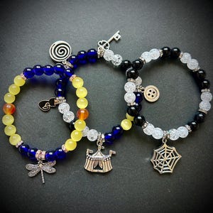 May include: Three beaded bracelets with charms. One bracelet has yellow and blue beads with a dragonfly charm. Another has yellow, blue, and clear beads with a circus tent charm. The third has black, white, and clear beads with a spiderweb charm.