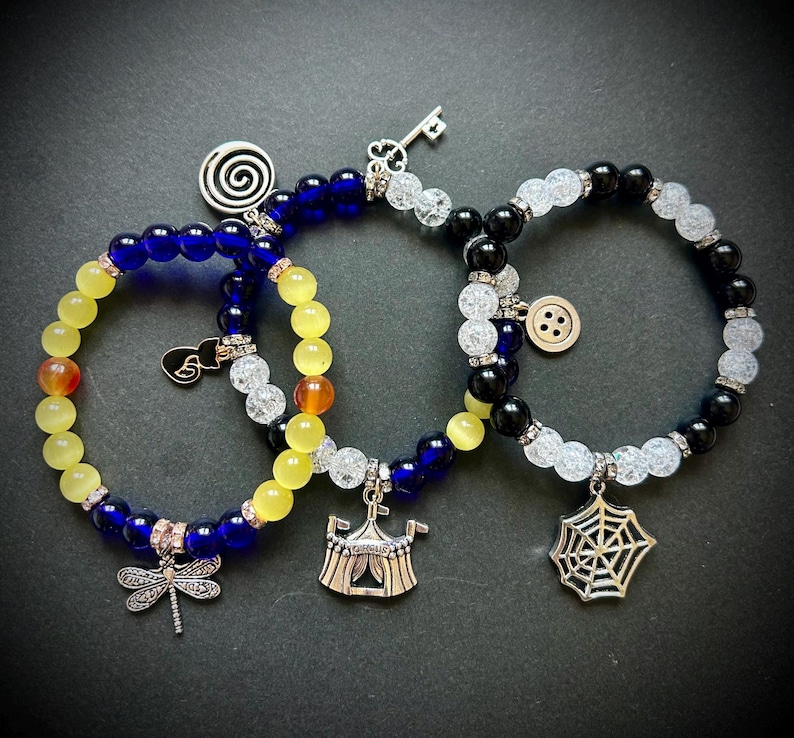 May include: Three beaded bracelets with charms. One bracelet has yellow and blue beads with a dragonfly charm. Another has yellow, blue, and clear beads with a circus tent charm. The third has black, white, and clear beads with a spiderweb charm.
