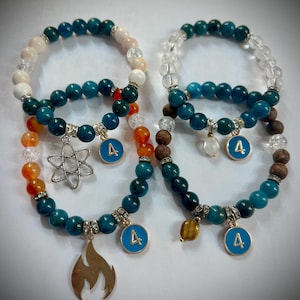 May include: Two beaded bracelets featuring teal, white, orange, and clear beads. Each bracelet has a silver charm, a blue charm with the number 4, and a gold flame charm. The bracelets are displayed on a white background.