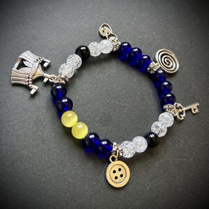 May include: A beaded bracelet with blue, yellow, black, and clear beads. The bracelet features silver charms including a key, a button, a spiral, and a decorative hat. The bracelet is on a dark background.