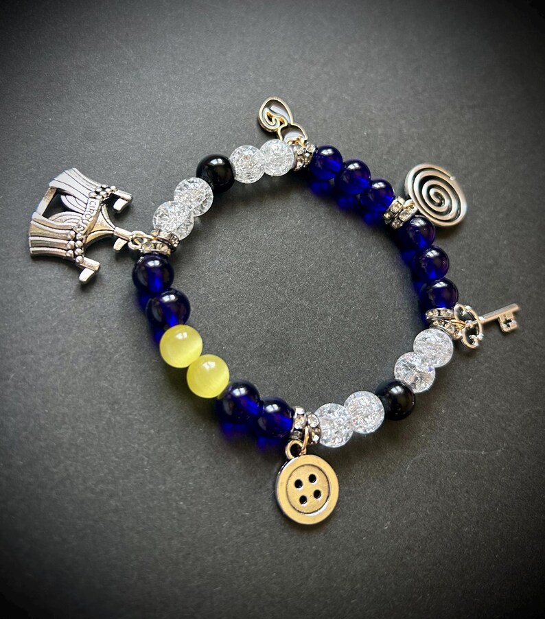 May include: A beaded bracelet with blue, yellow, black, and clear beads. The bracelet features silver charms including a key, a button, a spiral, and a decorative hat. The bracelet is on a dark background.
