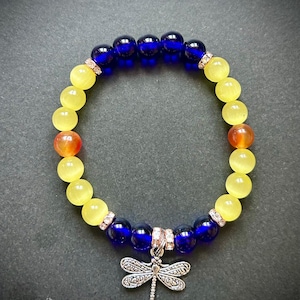 May include: A beaded bracelet featuring yellow, orange, and dark blue beads. The bracelet has a silver dragonfly charm. The beads are round and smooth. The bracelet is on a dark background.