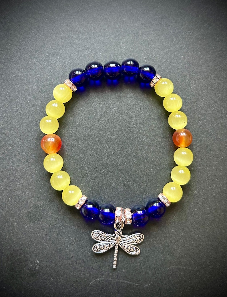 May include: A beaded bracelet featuring yellow, orange, and dark blue beads. The bracelet has a silver dragonfly charm. The beads are round and smooth. The bracelet is on a dark background.