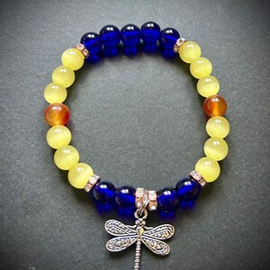 May include: A beaded bracelet featuring alternating yellow and blue beads, with a dragonfly charm. The bracelet includes orange and clear crystal accents. The dragonfly charm is silver-toned.