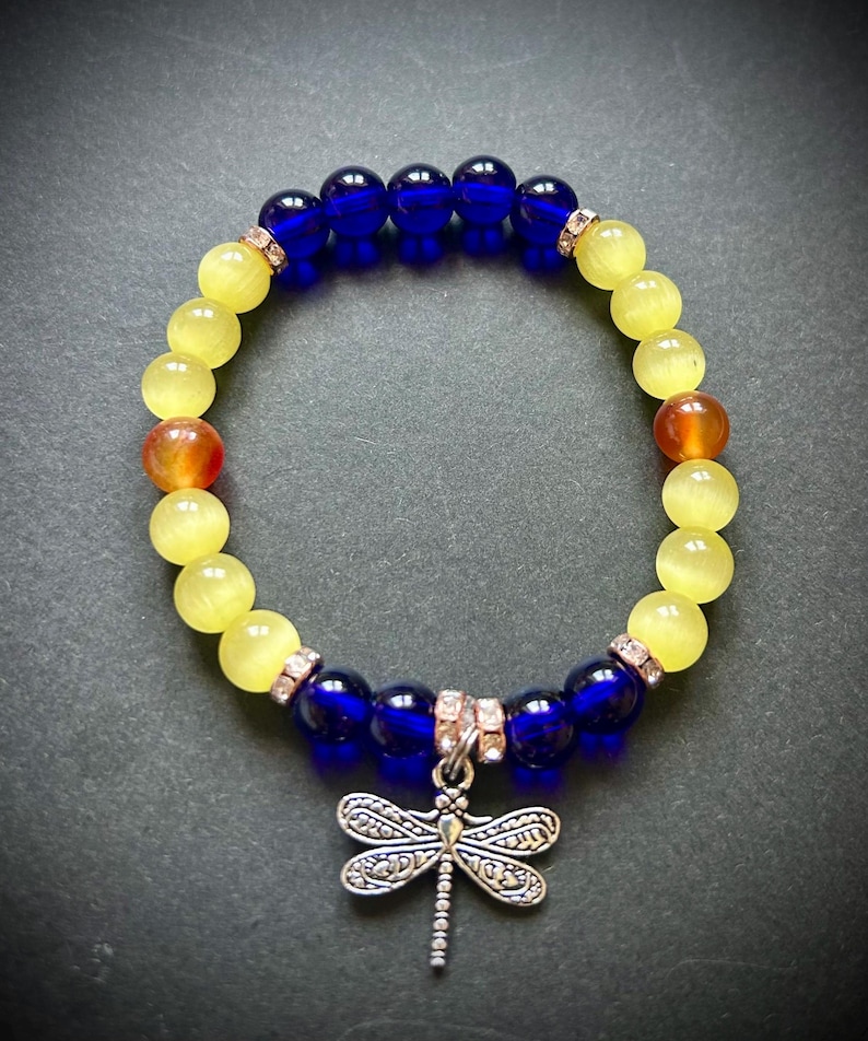 May include: A beaded bracelet featuring alternating yellow and blue beads, with a dragonfly charm. The bracelet includes orange and clear crystal accents. The dragonfly charm is silver-toned.