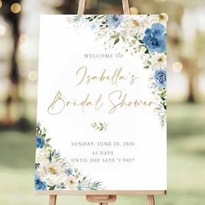 May include: A white sign on a wooden easel, decorated with blue and white floral arrangements. The sign reads "WELCOME TO Isabella's Bridal Shower" in gold script, with the date, "SUNDAY, JUNE 20, 2026," and the countdown "45 DAYS UNTIL SHE SAYS 'I DO!'"