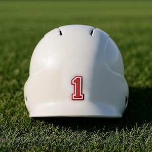 May include: A white baseball helmet with a red number 1 on the back. The helmet is resting on green grass. The helmet has several ventilation holes.