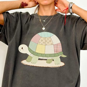 May include: Dark grey t-shirt featuring a cartoon turtle with a patchwork shell design in pastel colours. The turtle is on a beige base. The shirt has short sleeves and a relaxed fit.