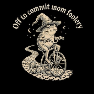 May include: Black graphic tee featuring a whimsical illustration of a frog riding a tricycle and wearing a wizard hat. The text "Off to commit mom foolery" is arched above the frog. The frog is riding on a cobblestone path.