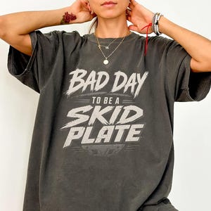 Könnte beinhalten: Dunkelgraues T-Shirt mit dem weißen Text "BAD DAY TO BE A SKID PLATE". Das Shirt hat kurze Ärmel und eine lockere Passform, geeignet für den täglichen Gebrauch.