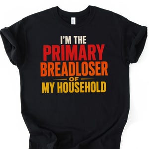 Op de afbeelding: Zwart T-shirt met ronde hals met de tekst "I'M THE PRIMARY BREADLOSER OF MY HOUSEHOLD" in wit, rood, oranje en geel. Het shirt heeft opgerolde mouwen en wordt gepresenteerd op een witte achtergrond.