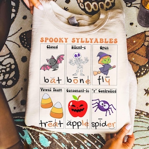 May include: A beige sweatshirt with a Halloween-themed graphic. The graphic features the text "SPOOKY SYLLYABLES" and illustrations of a bat, skeleton, witch, candy corn, apple, and spider. The sweatshirt is folded.