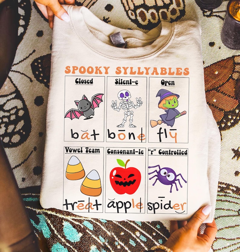 May include: A beige sweatshirt with a Halloween-themed graphic. The graphic features the text "SPOOKY SYLLYABLES" and illustrations of a bat, skeleton, witch, candy corn, apple, and spider. The sweatshirt is folded.