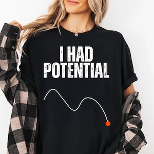 Può includere: T-shirt nero con la scritta bianca "I HAD POTENTIAL" e un grafico a linee bianche con un punto arancione. La persona indossa una camicia a quadri bianca e nera sopra la t-shirt.