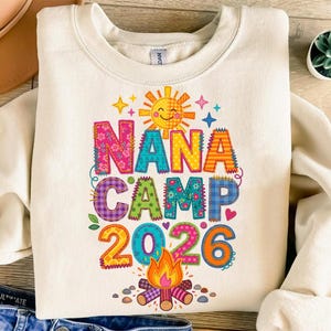 May include: A cream-colored crewneck sweatshirt featuring a colorful graphic print with the text NANA CAMP 2026. The design includes a smiling sun, a campfire, and letters decorated with various patterns like floral, plaid, and polka dots.
