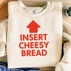 May include: Cream-colored sweatshirt with the text "INSERT CHEESY BREAD" in red, topped with a red arrow pointing upwards. The sweatshirt is folded, with a small succulent plant visible in the background.