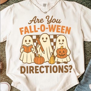 May include: Cream-colored t-shirt with the text "Are You FALL-O-WEEN DIRECTIONS?". The shirt features three cartoon ghosts holding a book, a pencil, and a jack-o'-lantern. The design includes pumpkins and a checkered background, perfect for Halloween.