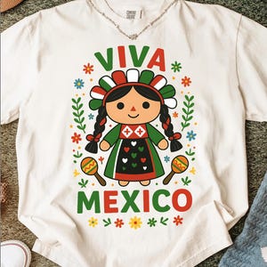 May include: White t-shirt with a colourful graphic featuring a Mexican doll design. The design includes the words "VIVA MEXICO" in red and green, surrounded by floral and leaf accents. The doll wears a traditional dress and holds maracas.