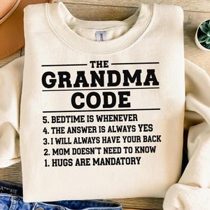 Op de afbeelding: Crèmekleurige sweatshirt met de tekst "THE GRANDMA CODE" in vette zwarte letters. Daaronder staan vijf regels, waaronder "HUGS ARE MANDATORY" en "THE ANSWER IS ALWAYS YES". De sweatshirt ligt op een houten oppervlak.