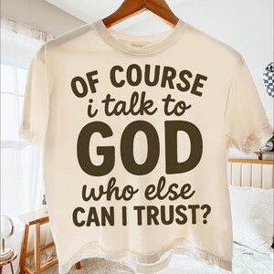 May include: Cream-colored t-shirt with the text "OF COURSE I talk to GOD who else CAN I TRUST?" in dark brown lettering. The shirt is hanging on a wooden hanger.