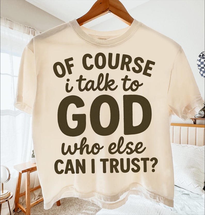 May include: Cream-colored t-shirt with the text "OF COURSE I talk to GOD who else CAN I TRUST?" in dark brown lettering. The shirt is hanging on a wooden hanger.
