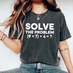 May include: Dark gray t-shirt with the text "SOLVE THE PROBLEM (9 x 7) + 4 = ?" in white. The shirt is a crew neck style and has a casual, worn look. The text is centered on the front of the shirt.