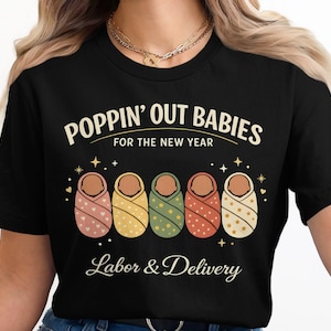 Op de afbeelding: Zwart T-shirt met de tekst "POPPIN' OUT BABIES FOR THE NEW YEAR" en de woorden "Labor & Delivery". Het ontwerp bevat vijf illustraties van ingebakerde baby's in roze, geel, groen, oranje en wit.