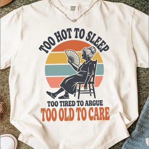 Puede incluir: Una camiseta de manga corta color blanco roto con un gráfico de una persona sentada en una silla usando un abanico. El texto dice: TOO HOT TO SLEEP, TOO TIRED TO ARGUE, TOO OLD TO CARE. El diseño incluye un fondo de atardecer circular en naranja, amarillo y azul verdoso.