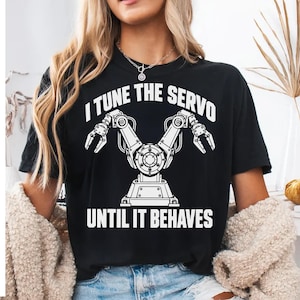 Op de afbeelding: Een zwart t-shirt met korte mouwen en een witte afbeelding van een robotarm met twee grijpers. De tekst "I TUNE THE SERVO" staat in het wit boven de afbeelding en de tekst "UNTIL IT BEHAVES" staat in het wit onder de afbeelding.