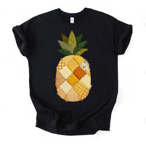 Può includere: Una t-shirt nera a maniche corte con una grande grafica di ananas sul davanti. L'ananas è realizzato con vari ritagli di tessuto trapuntato in tonalità giallo, arancione e crema, con foglie trapuntate verdi sulla parte superiore.