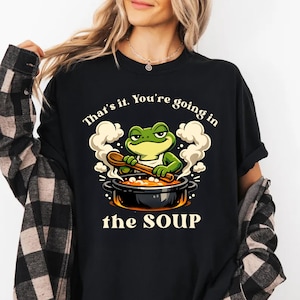 Puede incluir: Camiseta negra con un dibujo de un cocinero rana de dibujos animados removiendo sopa con una cuchara de madera. El texto encima de la rana dice "That's it. You're going in" y debajo "the SOUP".