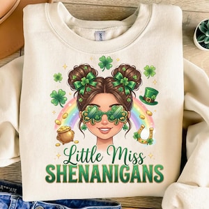 May include: Cream-colored sweatshirt with a St. Patrick's Day graphic. The graphic features a cartoon woman with shamrock glasses, a rainbow, and the text "Little Miss Shenanigans" in green. Other elements include a pot of gold and a horseshoe.