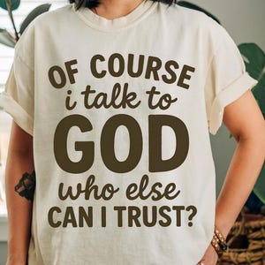 May include: Cream-colored t-shirt with the text "OF COURSE I talk to GOD who else CAN I TRUST?" in dark brown lettering. The shirt has rolled-up sleeves and a relaxed fit.