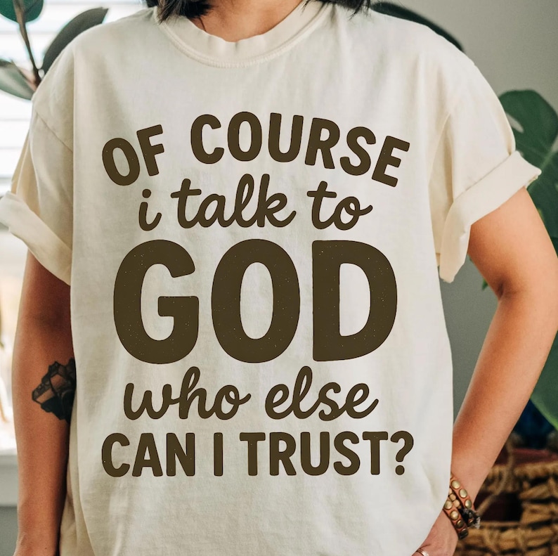 May include: Cream-colored t-shirt with the text "OF COURSE I talk to GOD who else CAN I TRUST?" in dark brown lettering. The shirt has rolled-up sleeves and a relaxed fit.