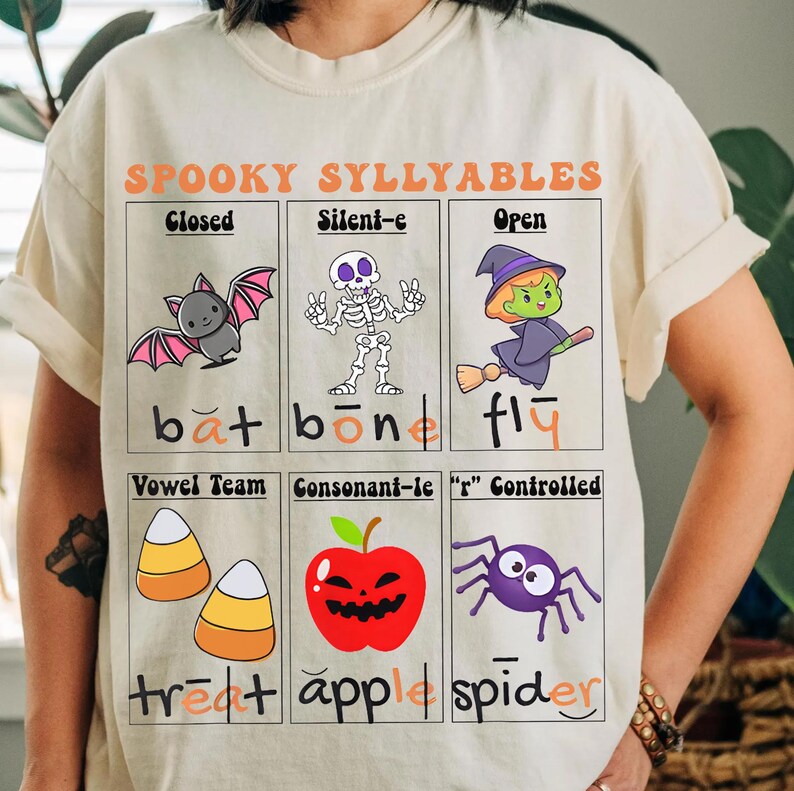 May include: Cream-colored t-shirt with a Halloween-themed graphic. The design features illustrations of a bat, skeleton, witch, candy corn, apple, and spider, along with the words "SPOOKY SYLLYABLES".