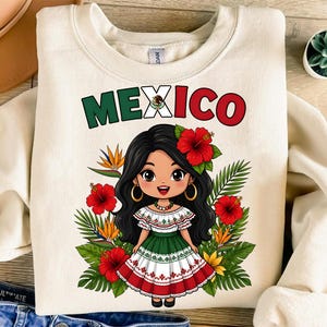 Pode incluir: Camisola creme com uma ilustração de desenho animado de uma menina com um vestido tradicional mexicano, rodeada por flores e folhagens. A palavra "MEXICO" está impressa no topo em verde e vermelho. O design apresenta cores vibrantes e detalhes intrincados.