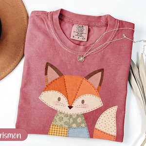 Może przedstawiać: Koszulka w kolorze karmazynowym z patchworkowym wzorem lisa. Lis ma pomarańczowe, brązowe i kremowe łaty. Srebrny naszyjnik z małą zawieszką spoczywa na koszulce. Koszulka jest oznaczona jako "Comfort Colors" i ma rozmiar S-P-CH.