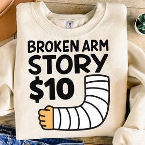 以下が含まれることがあります： 「BROKEN ARM STORY $10」の黒い文字が入ったクリーム色のスウェットシャツ。ギプスをはめた腕の漫画イラストがデザインされています。カジュアルで快適な衣服です。